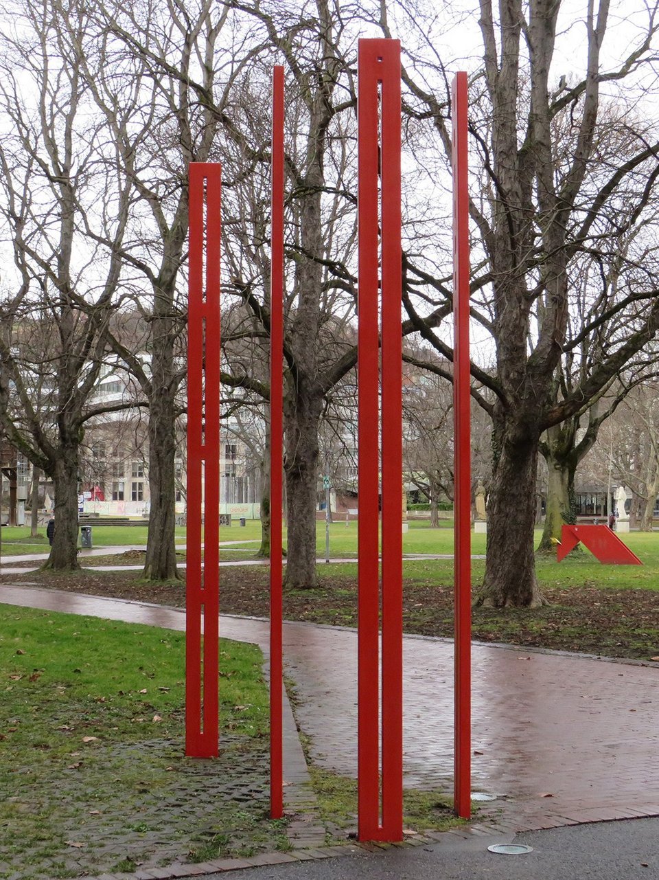 Four tall, narrow red steel steles in front of a park with large trees.