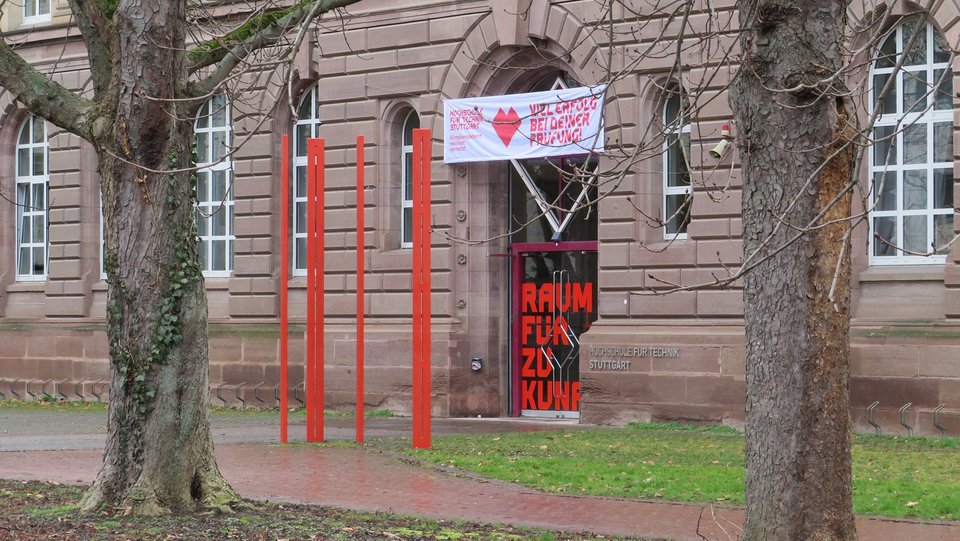 Four red steles in front of the entrance to the university building, photographed between two trees.